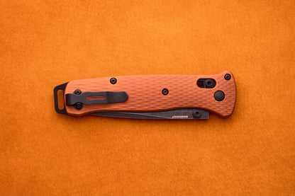 A folded Benchmade Bailout with an orange handle, textured grip, and black clip is set on an orange surface, highlighting its ultralight design ideal for everyday carry.