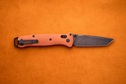 A Benchmade Bailout featuring a textured orange handle and a dark tanto-style CPM-Magnacut blade, inspired by ultralight design, shown on an orange background.