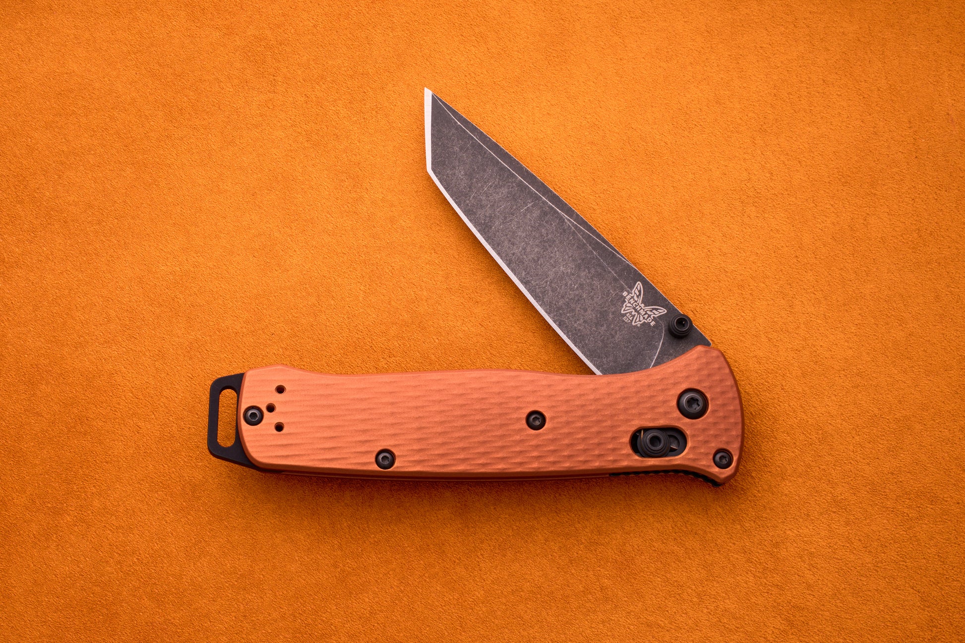 A partially open Benchmade Bailout ultralight knife with a textured orange handle and black clip, displayed on a bright orange surface. Its CPM-Magnacut blade features a stonewashed finish and the signature butterfly logo near the pivot, characteristic of the Bailout series.