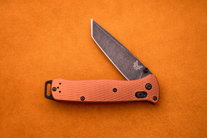 A partially open Benchmade Bailout ultralight knife with a textured orange handle and black clip, displayed on a bright orange surface. Its CPM-Magnacut blade features a stonewashed finish and the signature butterfly logo near the pivot, characteristic of the Bailout series.
