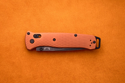 A closed Benchmade Bailout folding pocket knife with an orange textured handle rests on an orange surface, highlighting its ultralight design and the durability of its CPM-Magnacut blade.