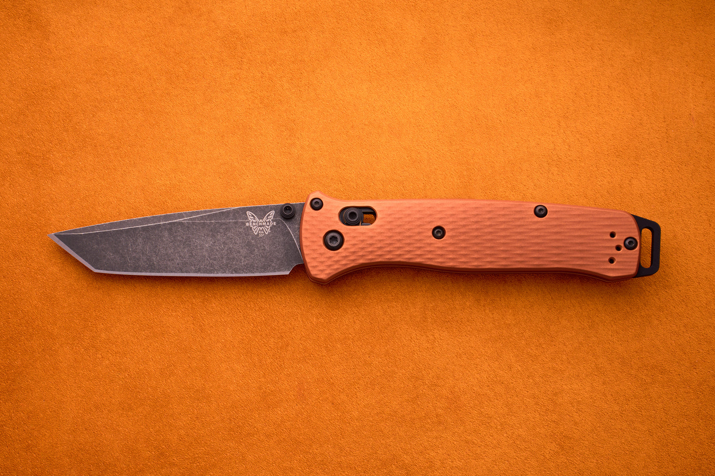 A Benchmade Bailout with a textured orange handle and a black CPM-Magnacut tanto blade is displayed on an orange surface, highlighting its ultralight, sleek design.