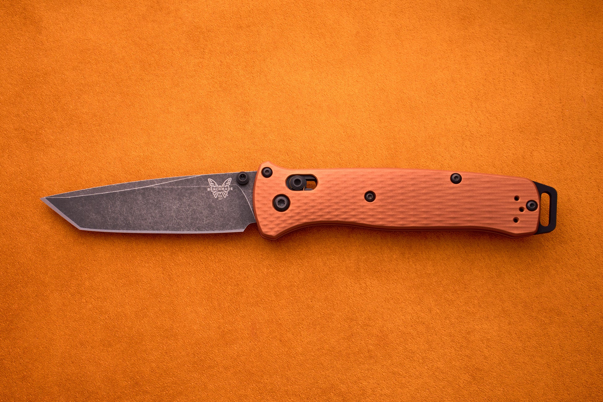 A Benchmade Bailout with a textured orange handle and a black CPM-Magnacut tanto blade is displayed on an orange surface, highlighting its ultralight, sleek design.