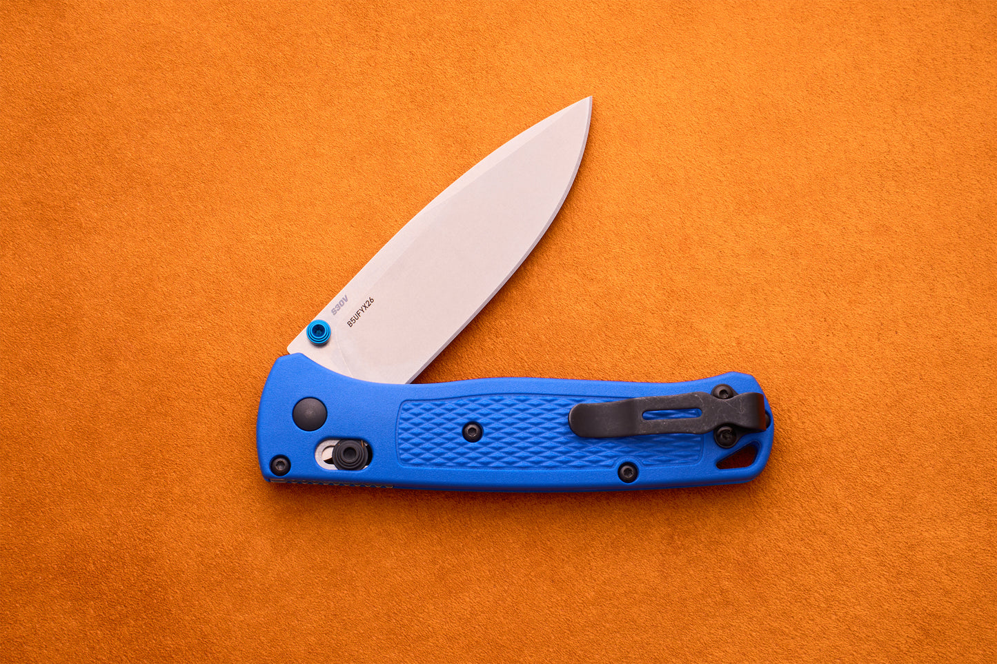 A Benchmade Bugout folding pocket knife with a blue textured handle and a partially open S30V stainless steel blade is resting on an orange surface.
