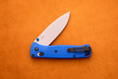 A Benchmade Bugout folding pocket knife with a blue textured handle and a partially open S30V stainless steel blade is resting on an orange surface.