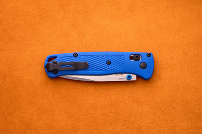 A Benchmade Bugout with a blue handle and a partially exposed S30V stainless steel blade rests on an orange textured surface, ready for your next outdoor adventure.