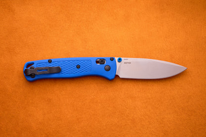 A Benchmade Bugout with a blue textured handle and a silver blade rests open on an orange background, perfect for your next outdoor adventure.