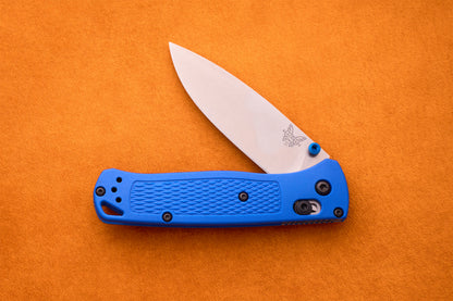 A Benchmade Bugout featuring a blue textured handle and a partially open silver blade rests on an orange surface—an ideal companion for outdoor adventures.