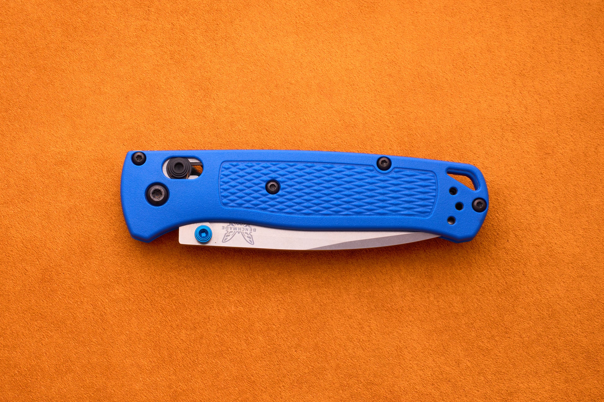 A closed Benchmade Bugout with a blue textured handle and a partially visible silver blade rests on an orange surface, making the Benchmade Bugout an excellent companion for any outdoor adventure.