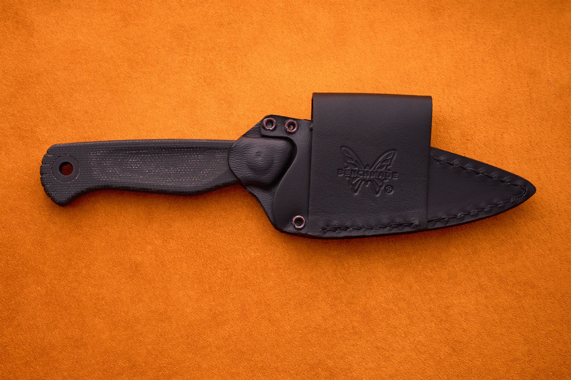 The Benchmade Dacian - Black Micarta is a compact fixed-blade knife crafted from CPM-Magnacut steel, featuring a textured black micarta handle and presented in a black leather sheath resting on an orange suede surface.