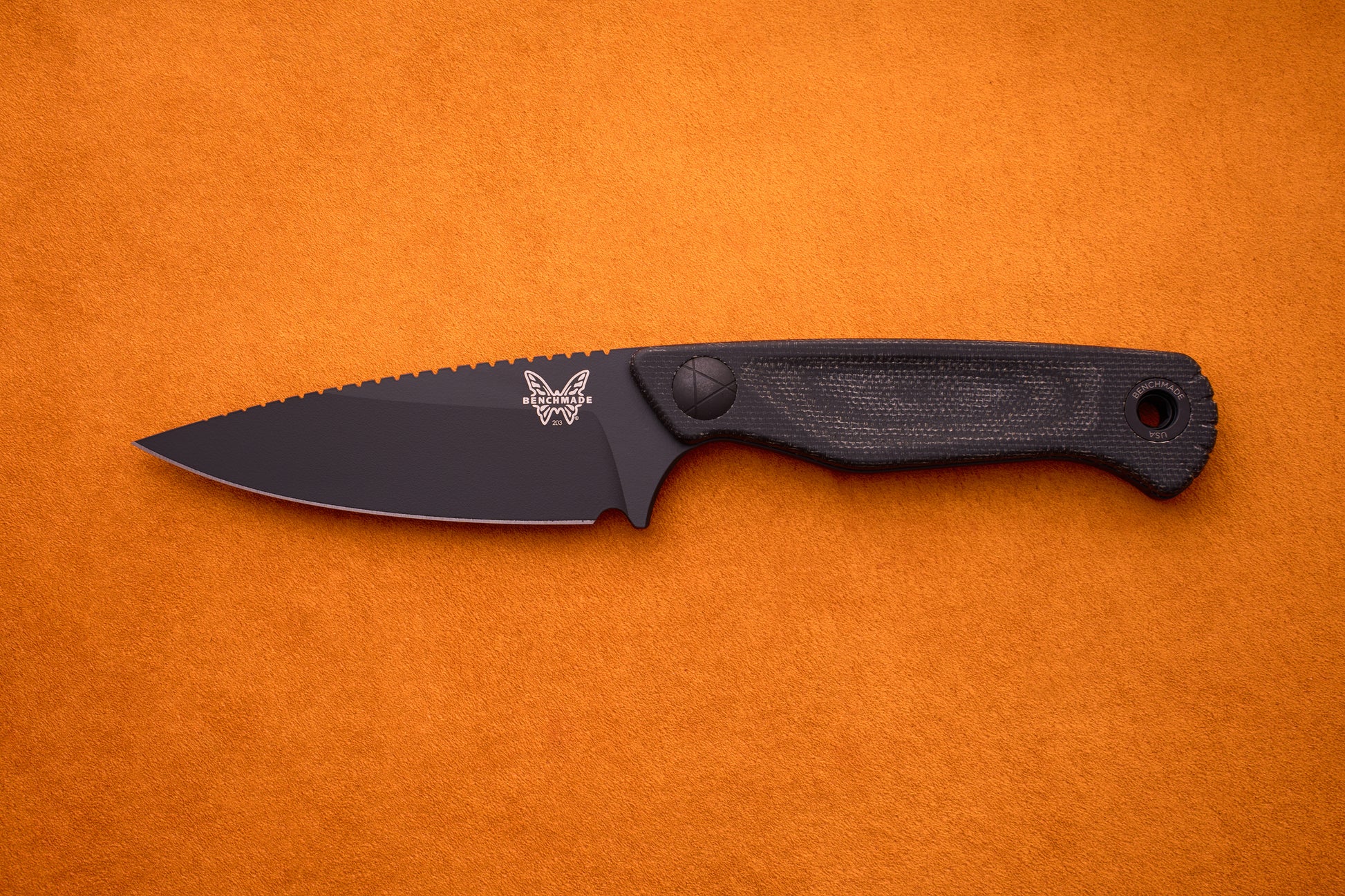 A compact fixed-blade Benchmade Dacian knife with a black Micarta handle and black CPM-Magnacut steel blade lies flat on an orange textured surface, featuring the butterfly logo near the base of the blade.