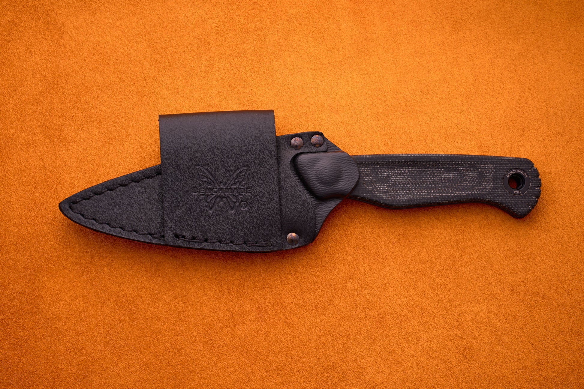 A Benchmade Dacian - Black Micarta compact fixed-blade knife with a black micarta handle and butterfly logo, shown in its fitted black sheath, is displayed on an orange textured surface.