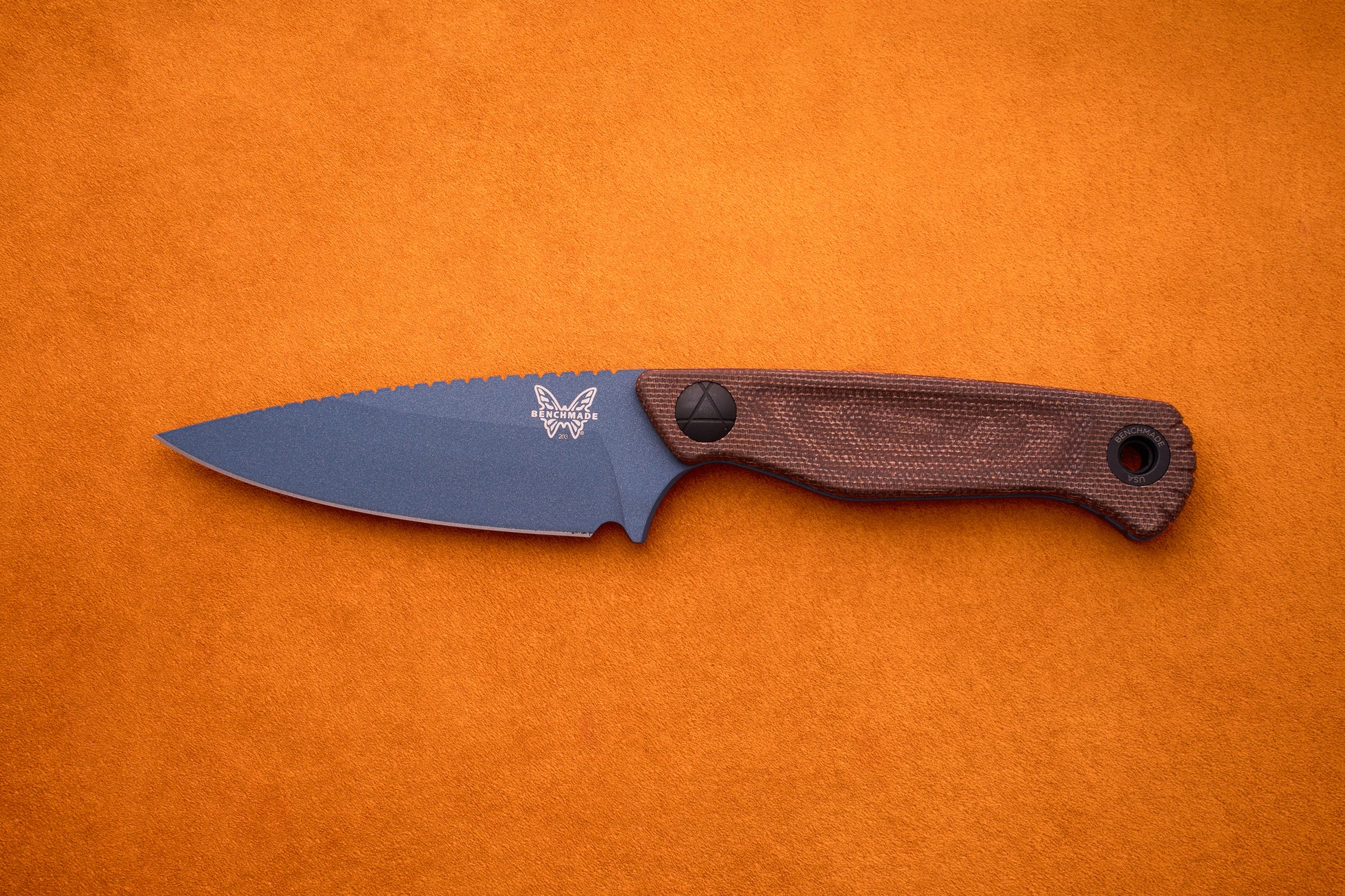 A Benchmade Dacian - Brown Micarta fixed-blade knife made from high-performance CPM-Magnacut steel, featuring a dark blue blade, a textured brown micarta handle, and a butterfly logo near the base of the blade, rests on an orange surface.