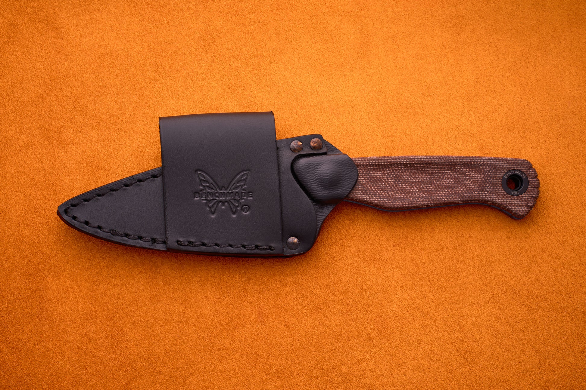 Benchmade Dacian - Brown Micarta: A fixed-blade knife crafted from premium CPM-Magnacut steel with a textured brown Micarta handle, presented in a black leather sheath embossed with the butterfly logo, shown resting on an orange surface.