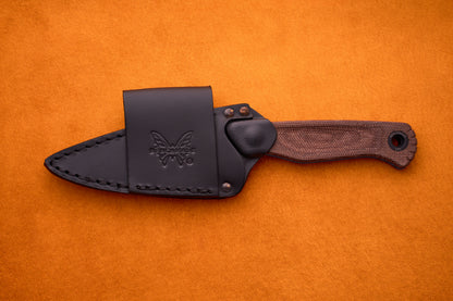 Benchmade Dacian - Brown Micarta: A fixed-blade knife crafted from premium CPM-Magnacut steel with a textured brown Micarta handle, presented in a black leather sheath embossed with the butterfly logo, shown resting on an orange surface.