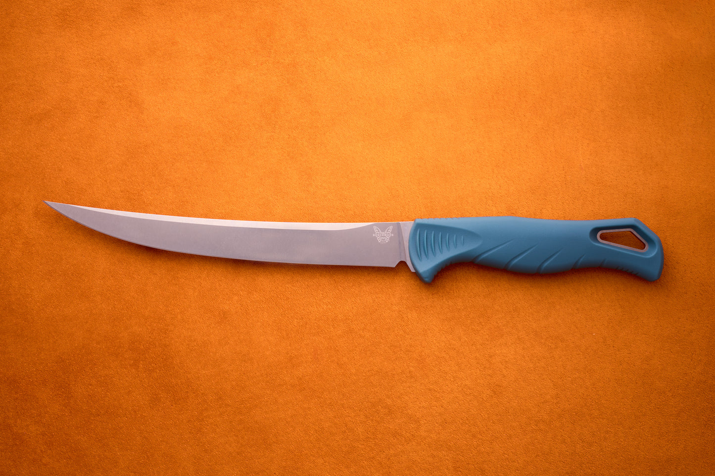 A Benchmade Fishcrafter—with its long, slender MagnaCut blade and blue ergonomic handle—rests on an orange textured surface, delivering expert filleting precision.