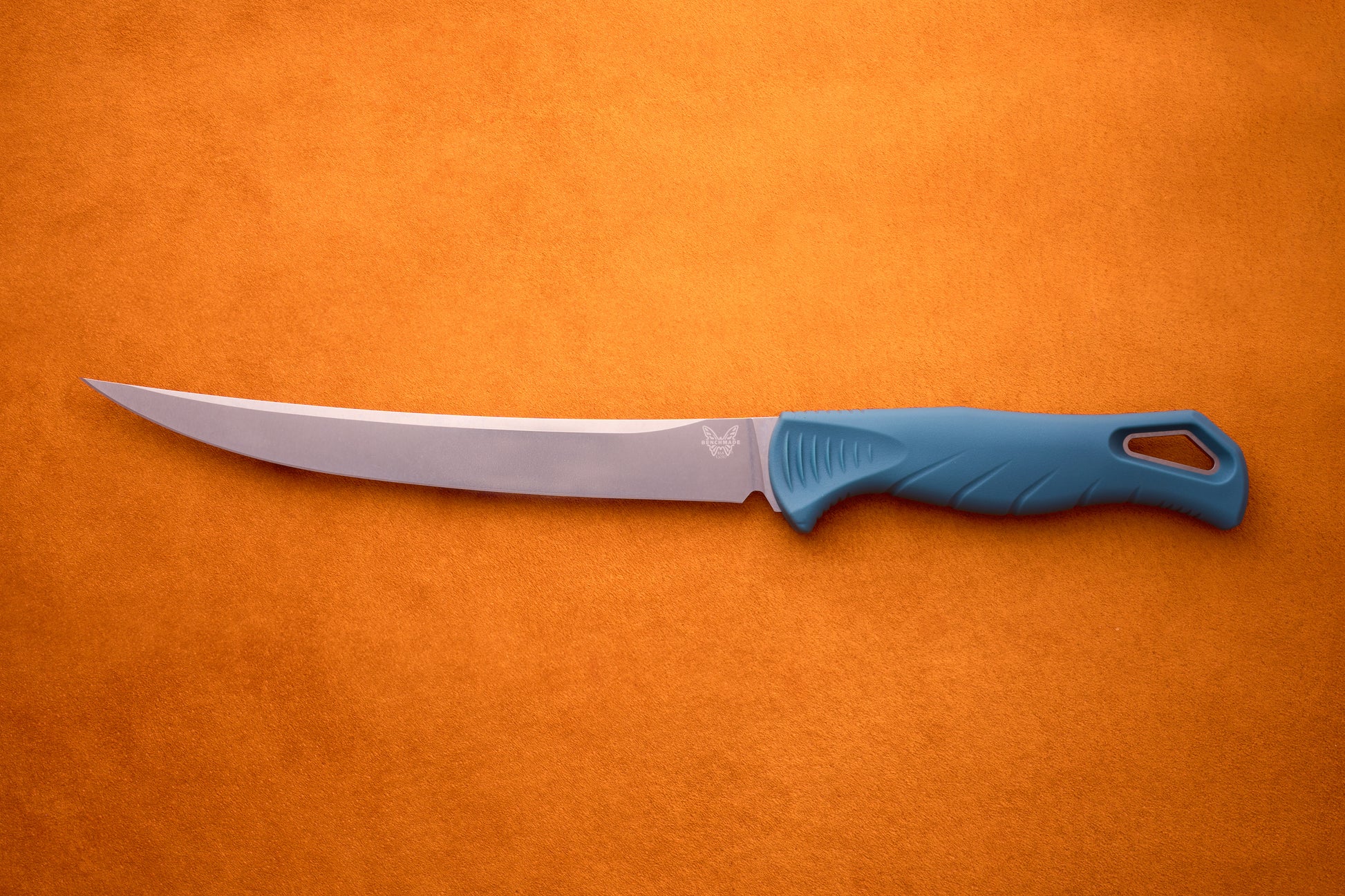 A Benchmade Fishcrafter—with its long, slender MagnaCut blade and blue ergonomic handle—rests on an orange textured surface, delivering expert filleting precision.