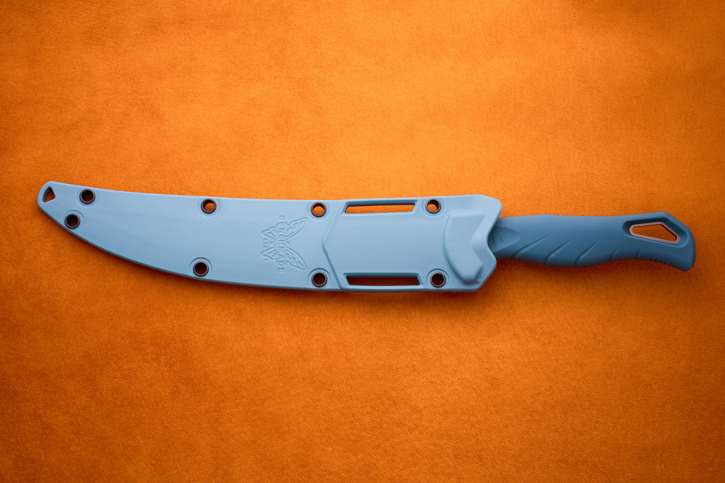 A blue Benchmade Fishcrafter knife with a protective sheath rests on an orange textured surface. The sheath, designed with multiple holes and adorned with a butterfly logo, securely covers the MagnaCut blade. The handle includes a lanyard hole and textured grip, making it perfect for precise filleting tasks.