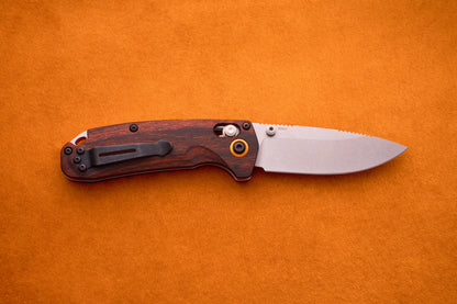 A Benchmade North Fork folding pocket knife with a wooden handle and a partially open CPM-S30V blade lies on an orange suede surface, showcasing its finely crafted design.