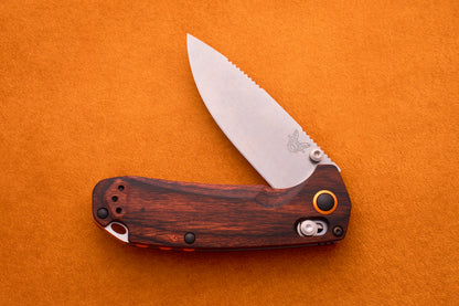 A Benchmade North Fork folding pocket knife featuring a wood-patterned handle and a partially open silver CPM-S30V blade, displayed on an orange textured background.