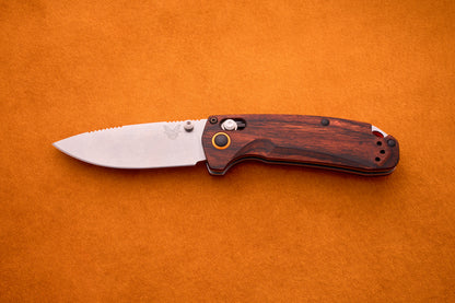 A Benchmade North Fork folding pocket knife, featuring a wood-grain handle and a razor-sharp CPM-S30V blade, lies open on an orange suede background, showcasing the craftsmanship of this premium hunting knife.