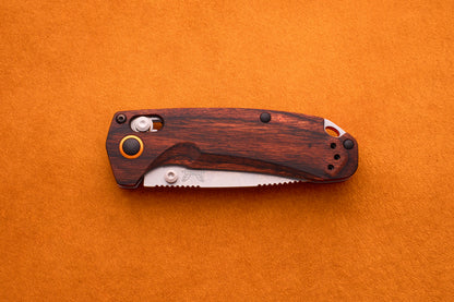 A Benchmade North Fork folding pocket knife with a wooden handle and a partially visible serrated CPM-S30V blade rests on an orange textured surface.