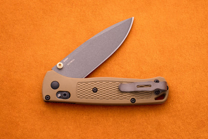 A tan-handled Benchmade Bugout folding pocket knife with a partially open blade is displayed on an orange background, highlighting its durable S30V stainless steel construction—an ideal choice for any outdoor adventure knife enthusiast.