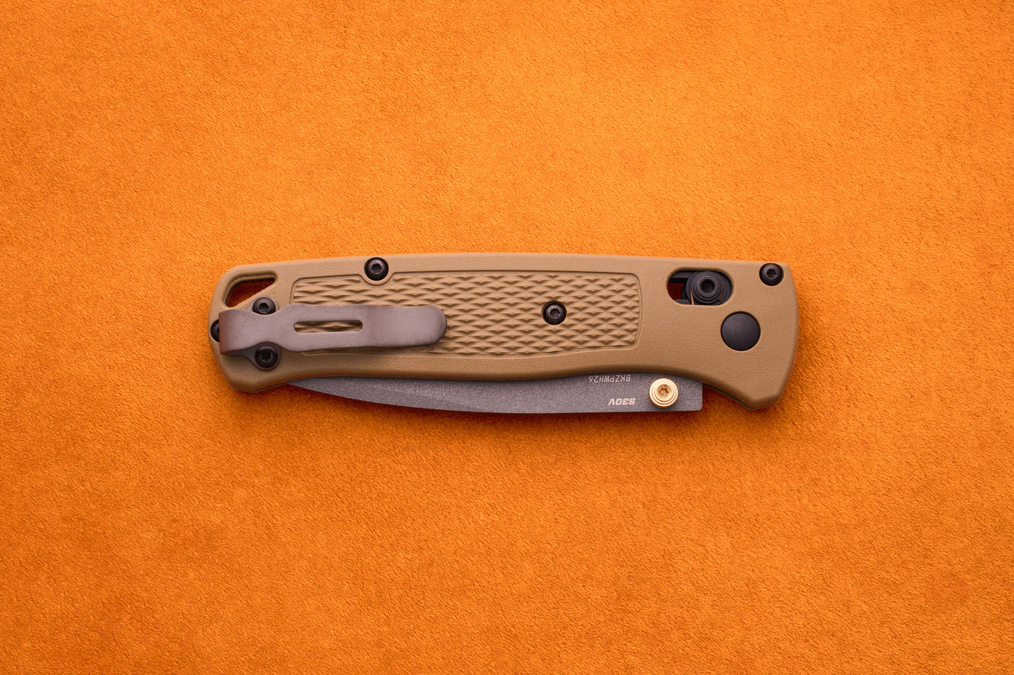 A closed Benchmade Bugout folding pocket knife with a tan textured handle rests on an orange surface, highlighting its reliable craftsmanship and durable S30V stainless steel—an ideal companion for any outdoor adventure knife enthusiast.