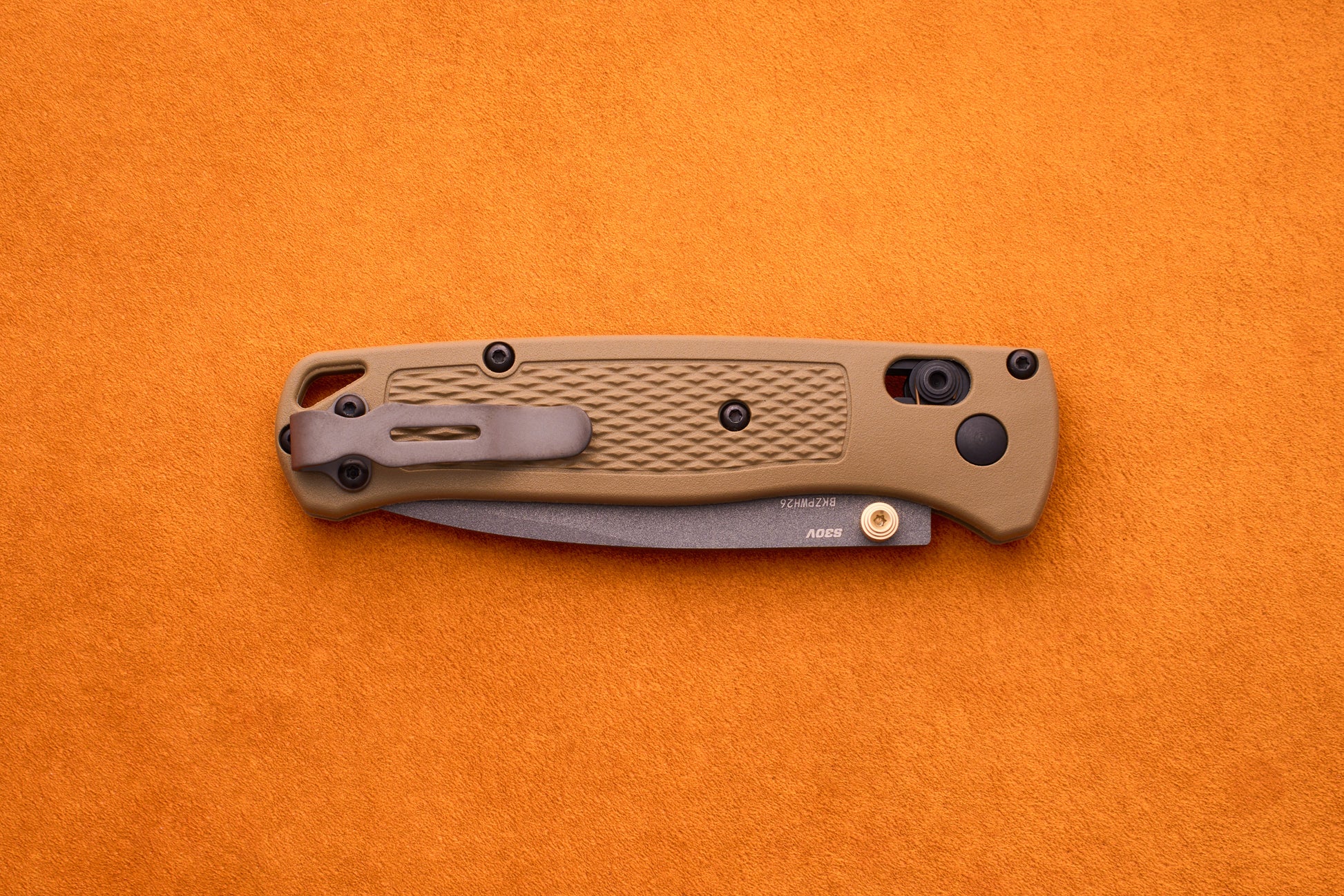 A closed Benchmade Bugout folding pocket knife with a tan textured handle rests on an orange surface, highlighting its reliable craftsmanship and durable S30V stainless steel—an ideal companion for any outdoor adventure knife enthusiast.