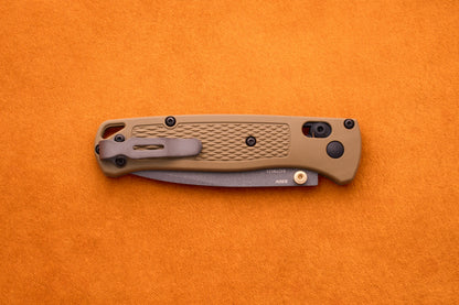 A closed Benchmade Bugout folding pocket knife with a tan textured handle rests on an orange surface, highlighting its reliable craftsmanship and durable S30V stainless steel—an ideal companion for any outdoor adventure knife enthusiast.