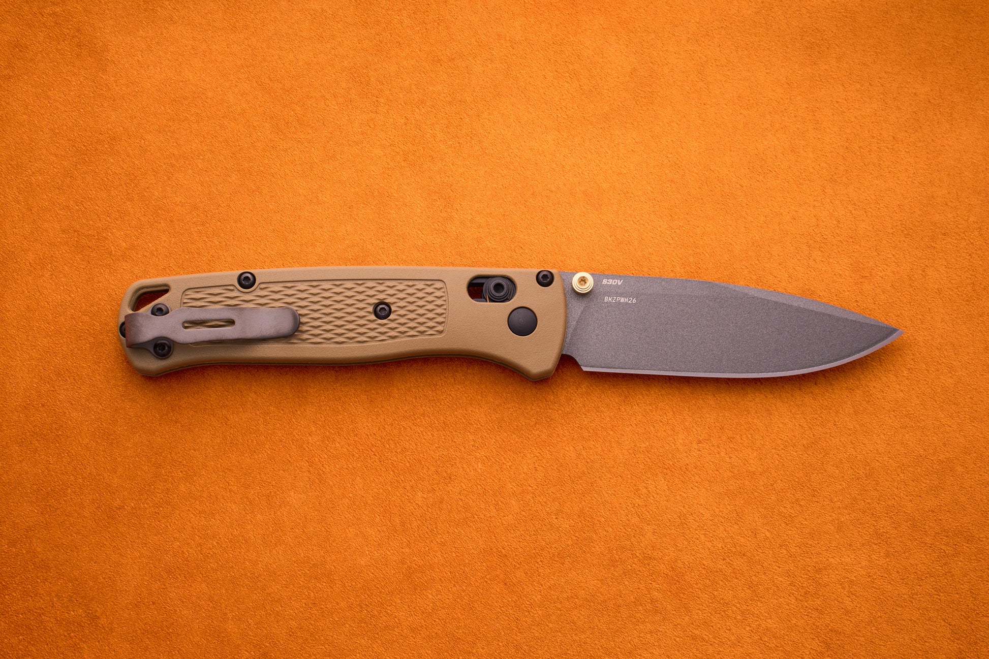 A Benchmade Bugout with a tan handle and gray blade is shown on an orange surface, highlighting it as a perfect knife for outdoor adventures.