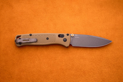 A Benchmade Bugout with a tan handle and gray blade is shown on an orange surface, highlighting it as a perfect knife for outdoor adventures.