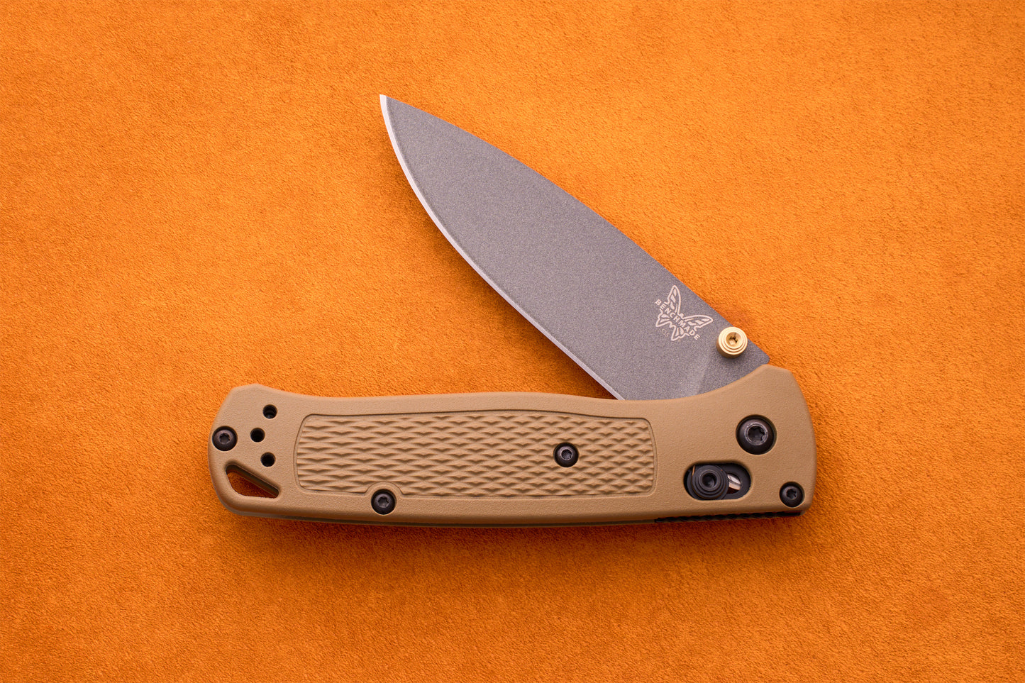 A Benchmade Bugout knife featuring a tan handle and a partially open silver blade, displayed on an orange textured surface—an ideal companion for outdoor adventures.
