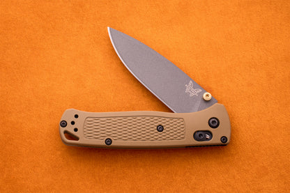 A Benchmade Bugout knife featuring a tan handle and a partially open silver blade, displayed on an orange textured surface—an ideal companion for outdoor adventures.
