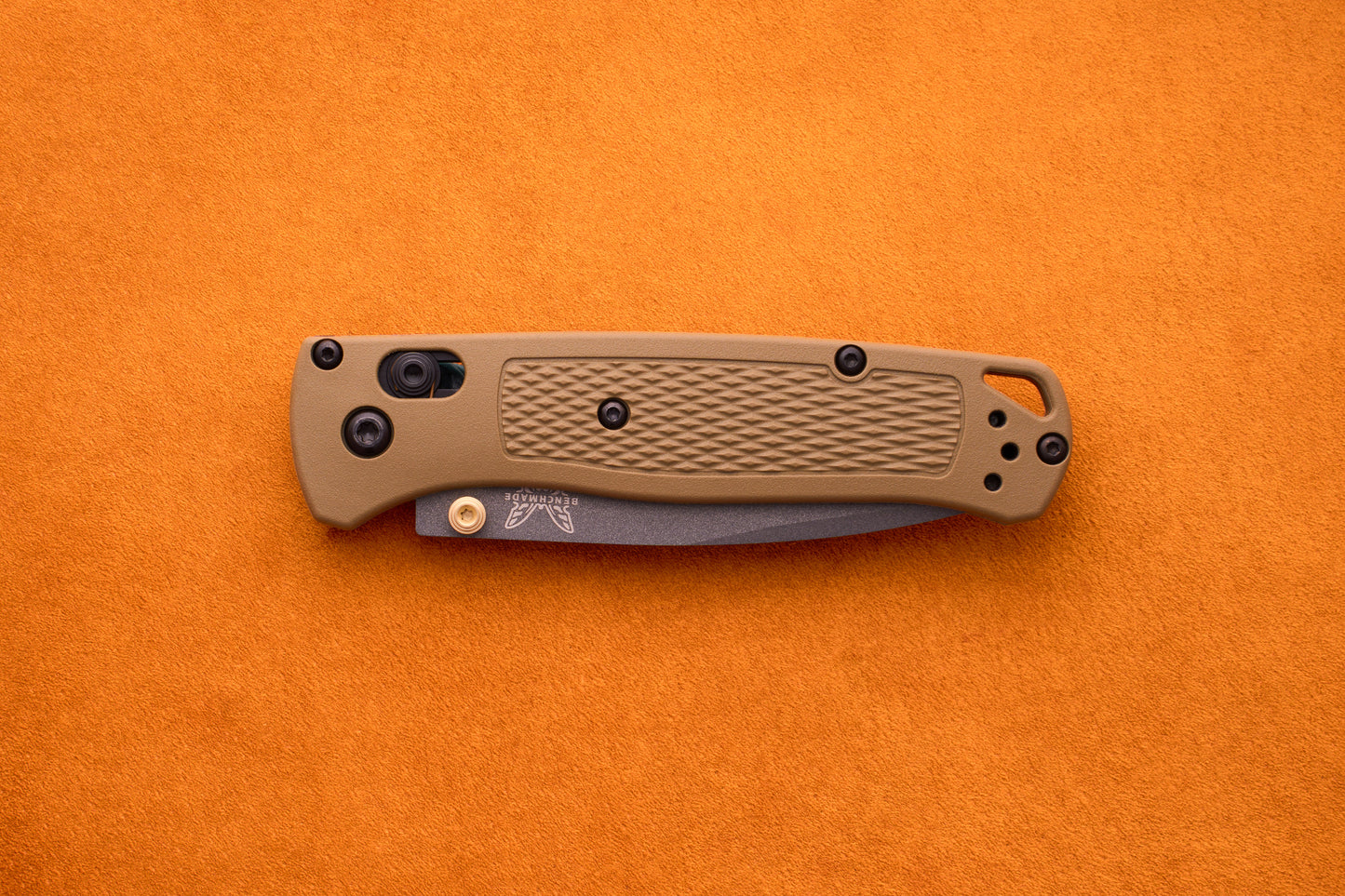 A folded Benchmade Bugout with a tan textured handle and black hardware rests on an orange surface, prepared for outdoor adventures.