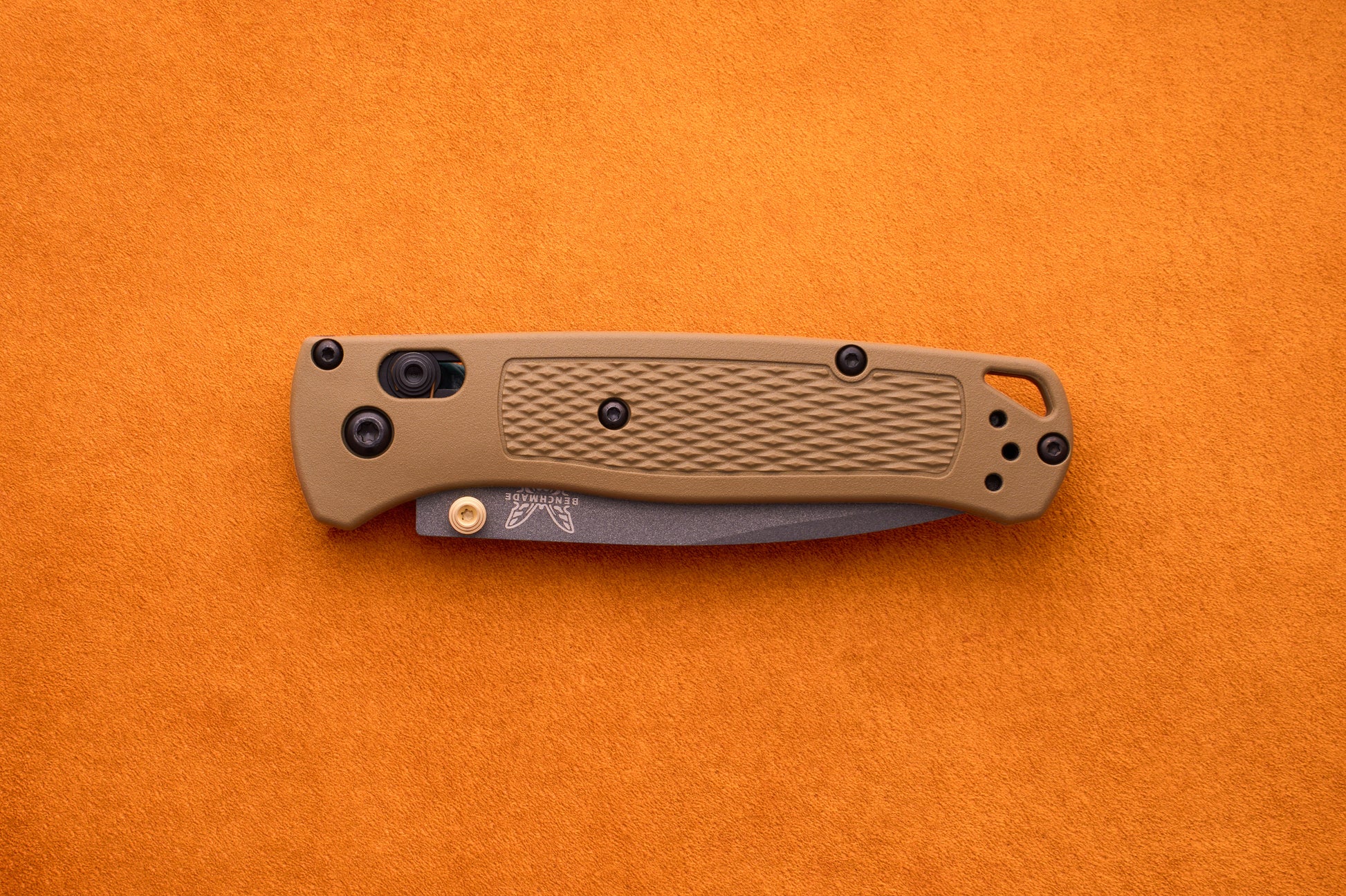 A folded Benchmade Bugout with a tan textured handle and black hardware rests on an orange surface, prepared for outdoor adventures.