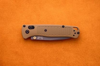 A folded Benchmade Bugout with a tan textured handle and black hardware rests on an orange surface, prepared for outdoor adventures.