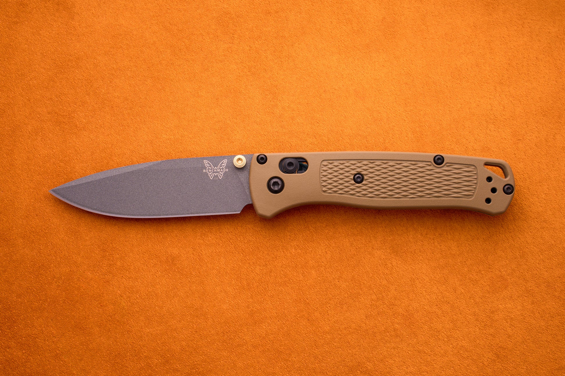 A Benchmade Bugout folding pocket knife with a brown textured handle and a gray S30V stainless steel blade is displayed on an orange surface, making it an ideal choice for outdoor adventures.