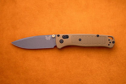 A Benchmade Bugout folding pocket knife with a brown textured handle and a gray S30V stainless steel blade is displayed on an orange surface, making it an ideal choice for outdoor adventures.