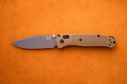 A Benchmade Bugout folding pocket knife with a brown textured handle and a gray S30V stainless steel blade is displayed on an orange surface, making it an ideal choice for outdoor adventures.
