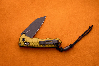 The Benchmade Full Immunity, featuring a textured yellow handle and a black M4 steel blade, is partially open and rests on an orange surface. A black cord with a bead is attached to the handle.
