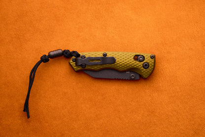 A Benchmade Full Immunity compact utility knife, featuring a textured green handle and black clip, is attached to a black paracord lanyard and set on an orange surface. Combining style and practicality, this knife reflects the renowned quality of Benchmade craftsmanship.