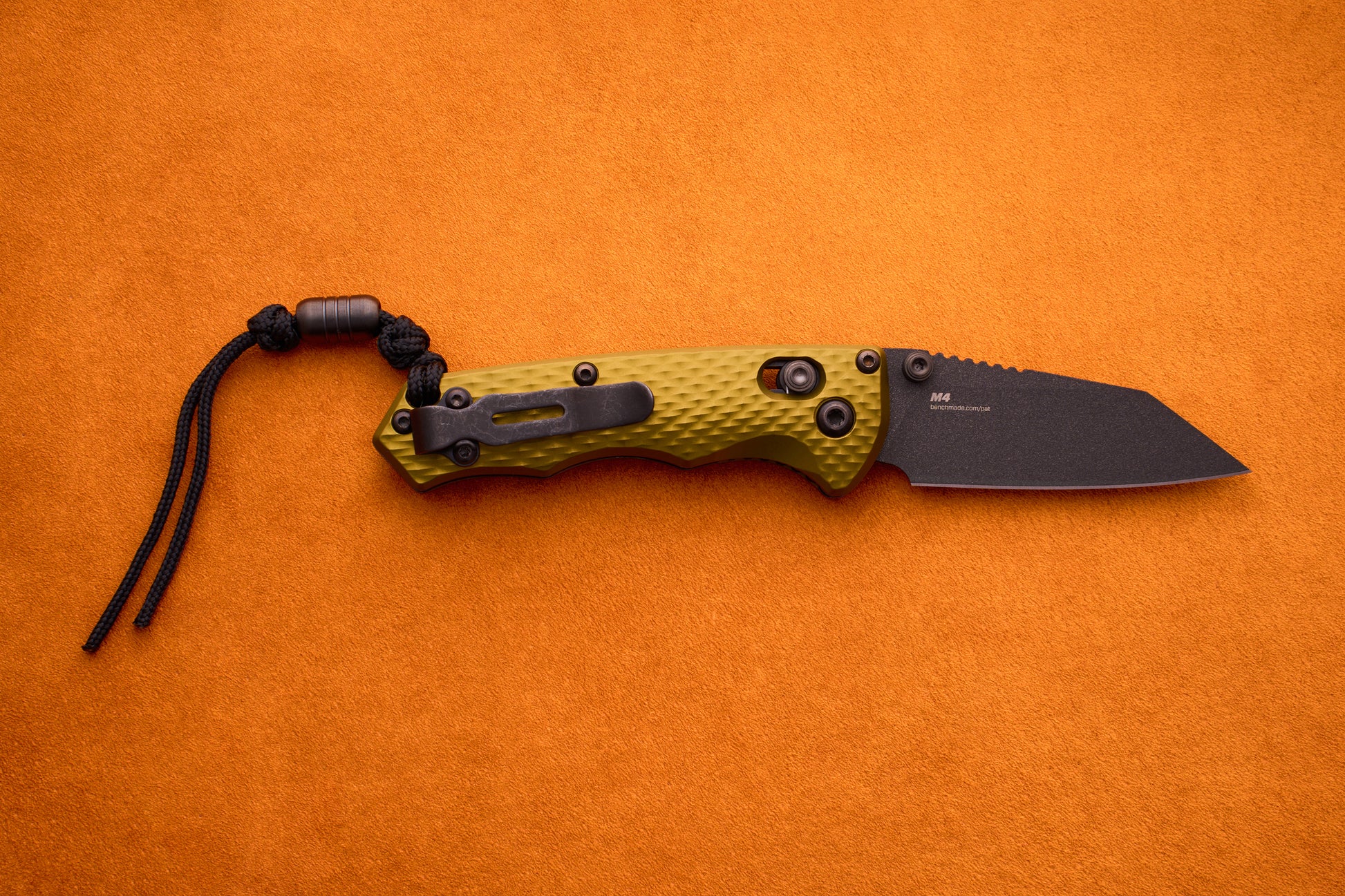A Benchmade Full Immunity folding pocket knife with a textured yellow-green handle, a black M4 steel blade, and an attached black paracord lanyard, displayed on an orange surface.