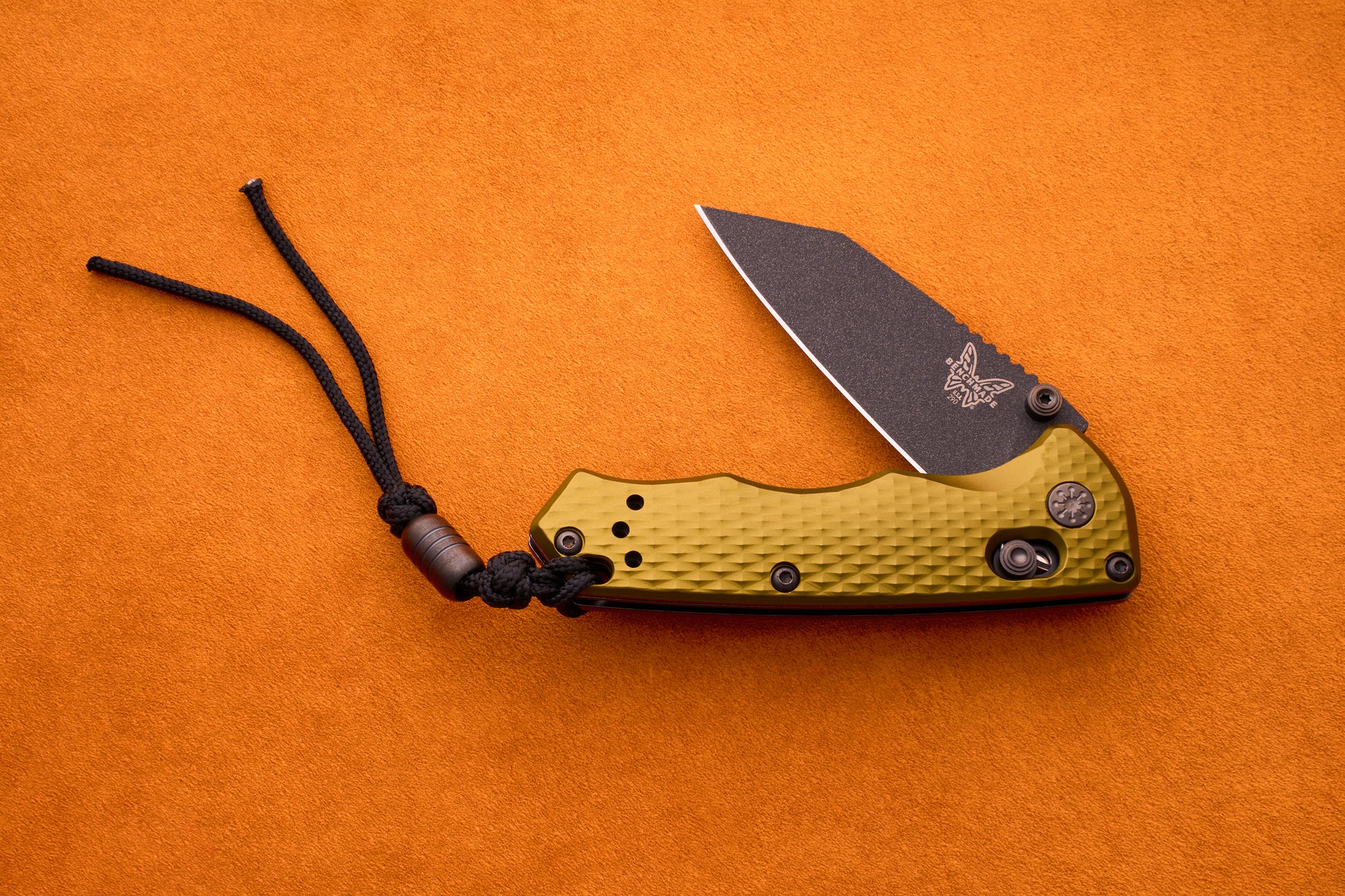 The Benchmade Full Immunity folding pocket knife features a compact design with a textured gold handle, a partially open black M4 steel blade, and an attached black lanyard. Resting on an orange surface, it blends durability and style in a sleek profile.