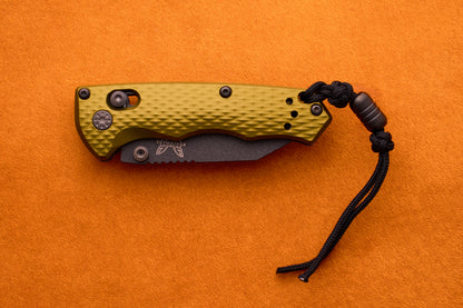 A closed Benchmade Full Immunity knife featuring a textured olive green handle and a black lanyard, made with robust M4 steel, rests on an orange surface.