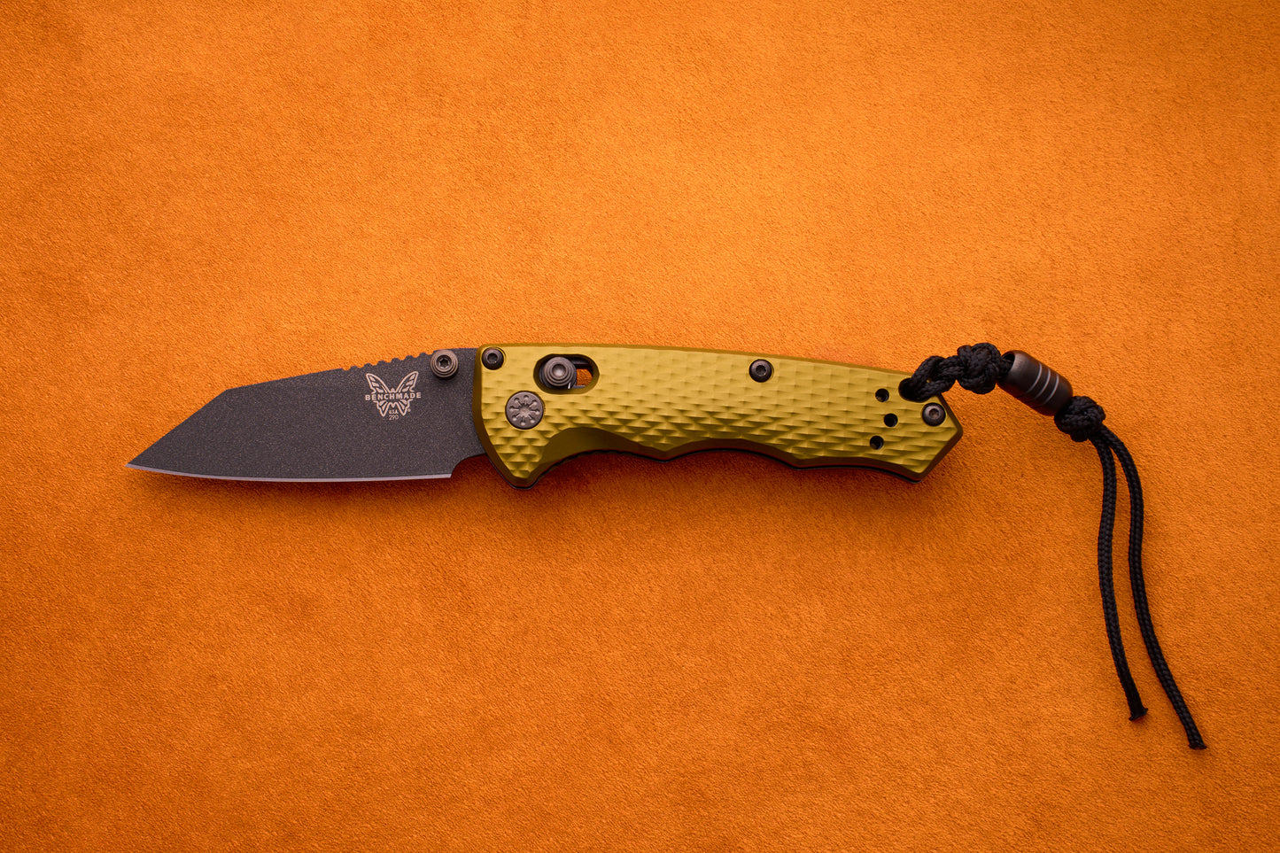 A Benchmade Full Immunity folding pocket knife, featuring a textured olive green handle, a black M4 steel blade, and a black cord lanyard—embodying signature rugged elegance—is displayed on an orange background.