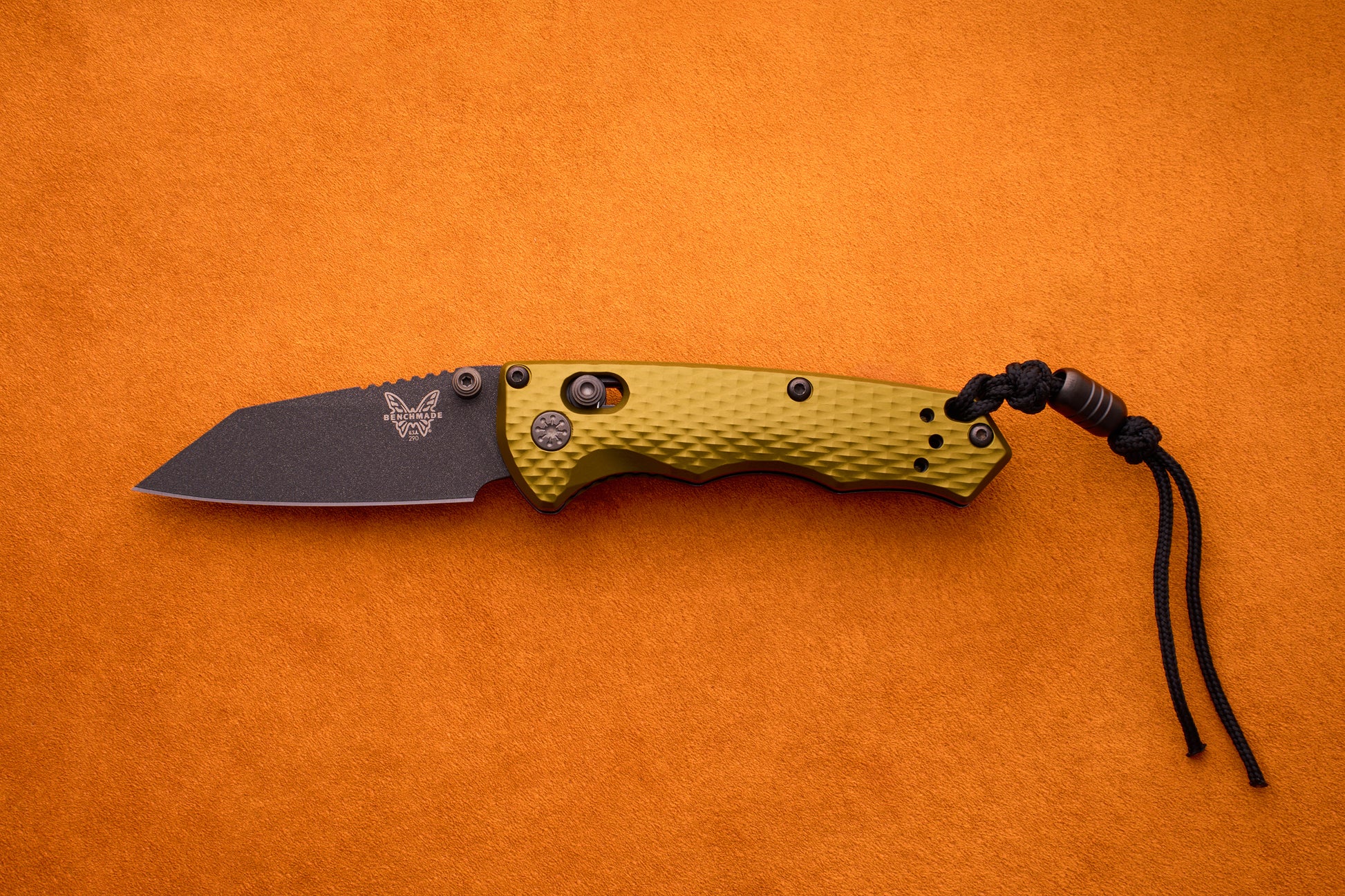 A Benchmade Full Immunity folding pocket knife, featuring a textured olive green handle, a black M4 steel blade, and a black cord lanyard—embodying signature rugged elegance—is displayed on an orange background.