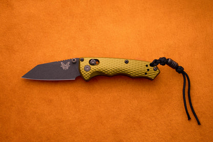 A Benchmade Full Immunity folding pocket knife, featuring a textured olive green handle, a black M4 steel blade, and a black cord lanyard—embodying signature rugged elegance—is displayed on an orange background.