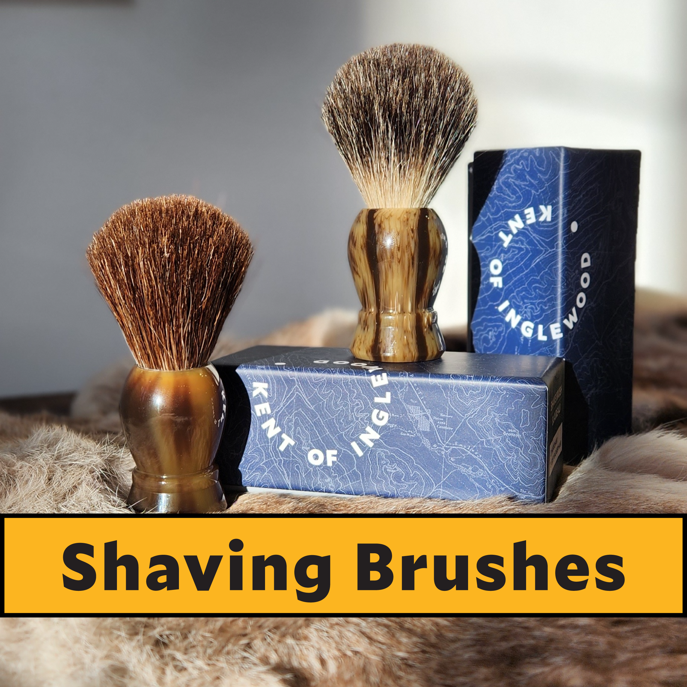 Two shaving brushes with wooden handles on a box labeled 'Kent of Inglewood' with a blurred background.
