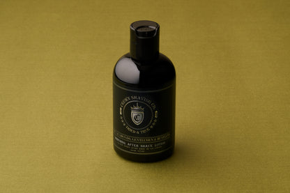 A black bottle of Crown Shaving Co. Aftershave Lotion sits on an olive green surface, its label facing forward and highlighting its reputation as a moisturizing balm ideal for sensitive skin.